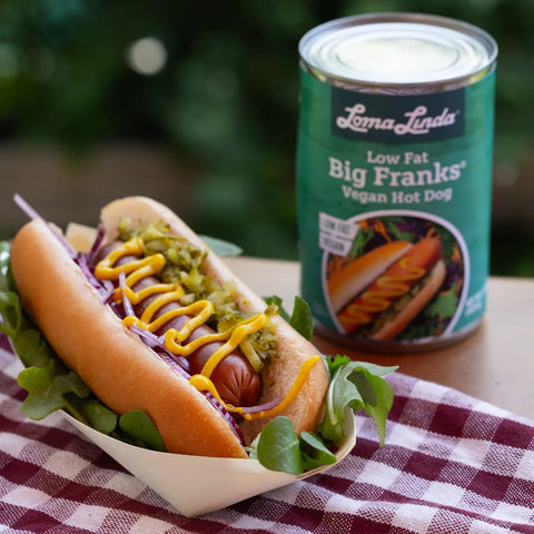 Single Loma Linda Big Frank on a bun with mustard, relish, and onions, served in a hot dog tray over a bed of arugula – plant-based hot dog alternative with classic toppings