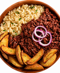 Overhead view of a wooden bowl filled with a meal consisting of couscous, UnMeat corned beef style, potato wedges, and red onion rings on a white background.