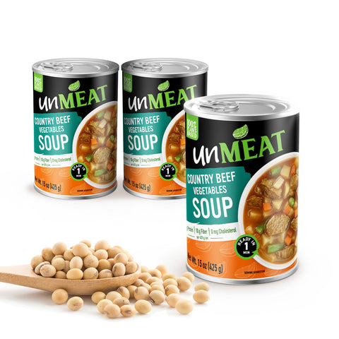 Three cans of 'unMEAT' country beef vegetables soup with a wooden spoonful of beans on a white background.